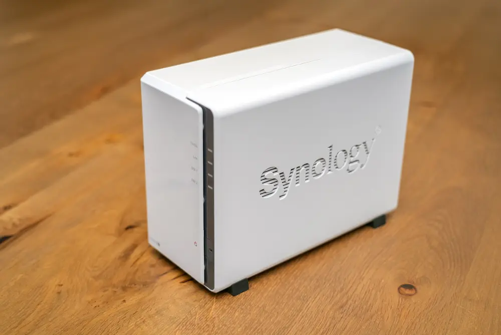 A small white Synology NAS with two drive bays, about the size of a shoebox, on a wooden table
